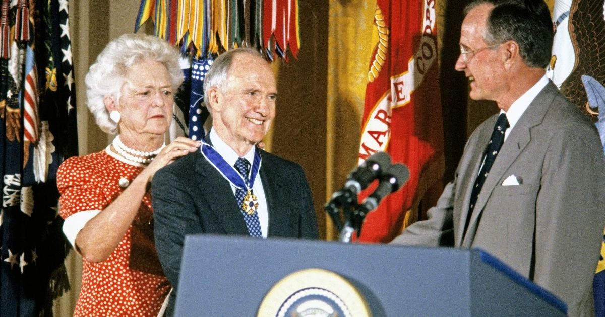 First Lady Barbara Bush fastens the Presidential Medal of Freedom around the neck of national security adviser Brent Scowcroft as he shakes hands with President George H.W. Bush during a ceremony in the White House's East Room in Washington, D.C., on July 3, 1991.