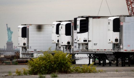 In this May 6, 2020, file photo, the Statue of Liberty is visible behind refrigerator trucks intended for storing corpses that are staged in a lot at the 39th Street pier, in the Brooklyn borough of New York.