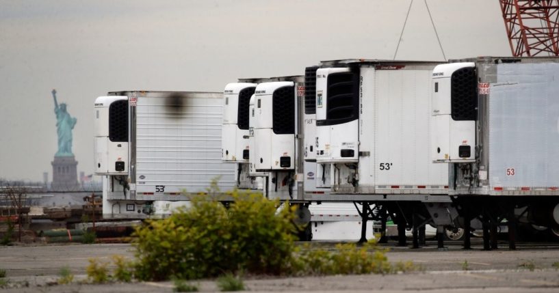 In this May 6, 2020, file photo, the Statue of Liberty is visible behind refrigerator trucks intended for storing corpses that are staged in a lot at the 39th Street pier, in the Brooklyn borough of New York.