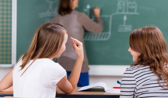 Two girls talk in class while a teacher writes on a blackboard.