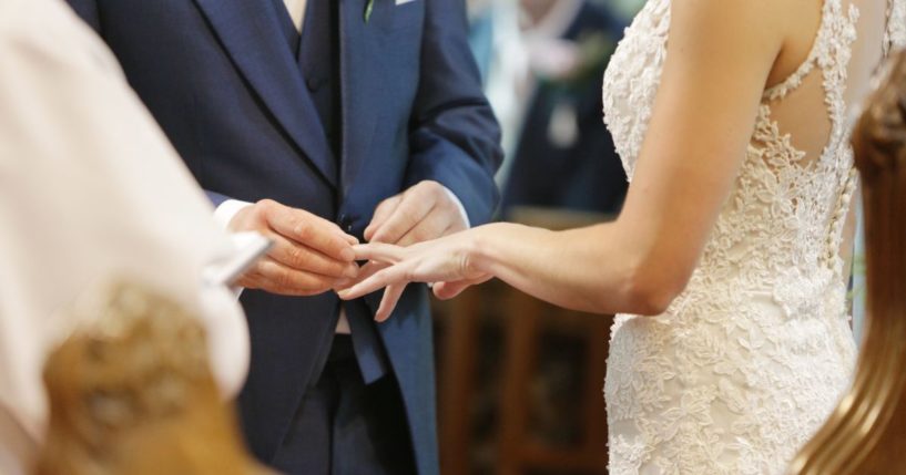 A groom places a ring on his bride's finger in this stock image.