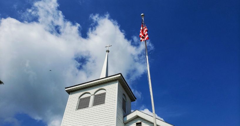 The American flag flies next to a church steeple in this stock image.