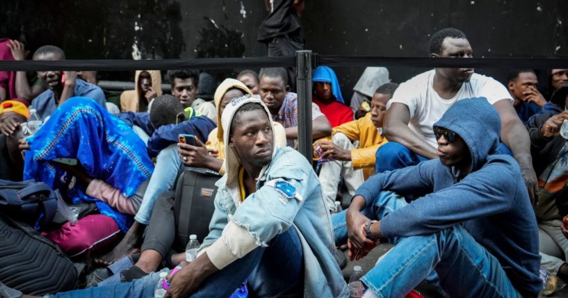 Migrants sit in a queue outside of The Roosevelt Hotel that is being used by the city as temporary housing, in a file photo from July 31 in New York City.