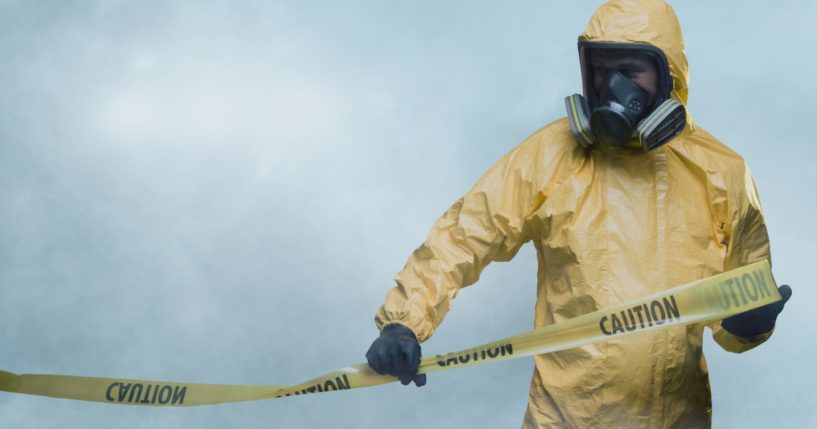 This stock image shows a worker in a hazmat suit putting up caution tape.