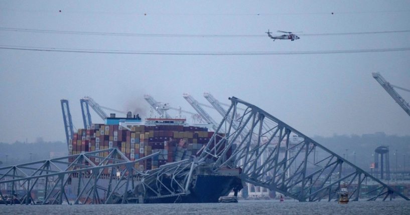 a helicopter flying over the container ship Dali as it rests against wreckage of the Francis Scott Key Bridge