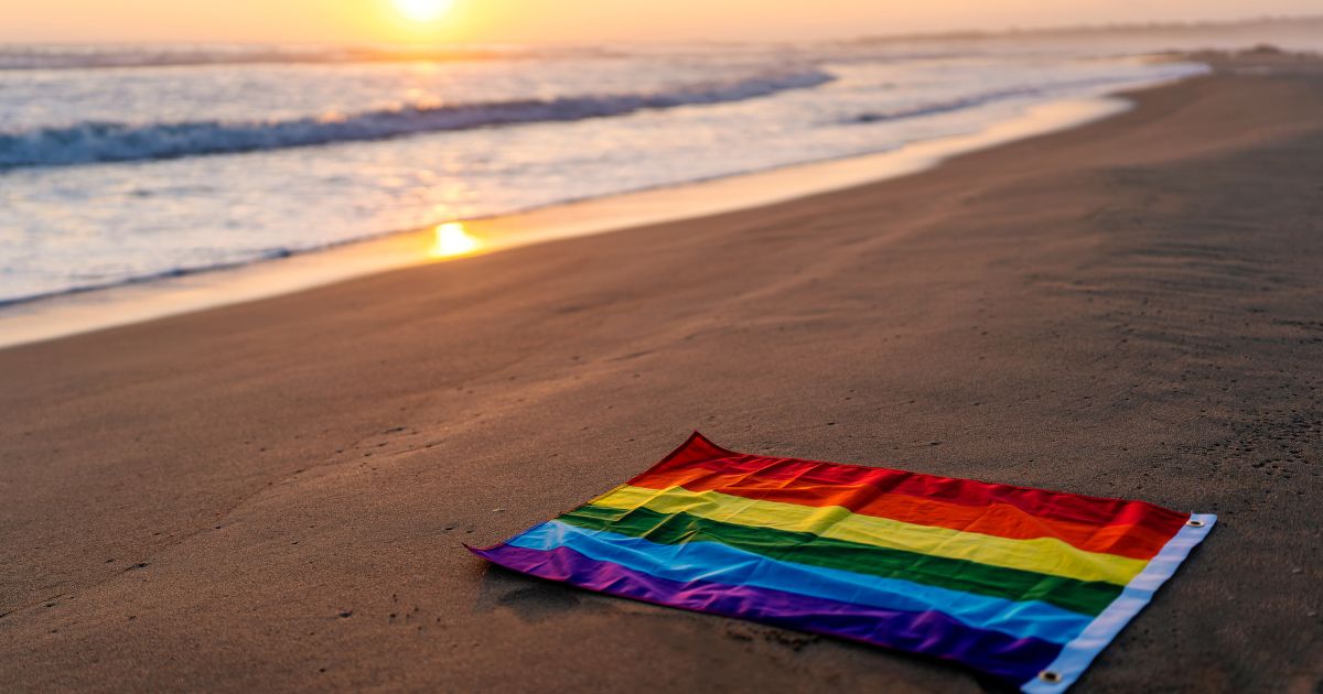 This image shows a rainbow 'pride' flag on the shore of the beach at sunset.