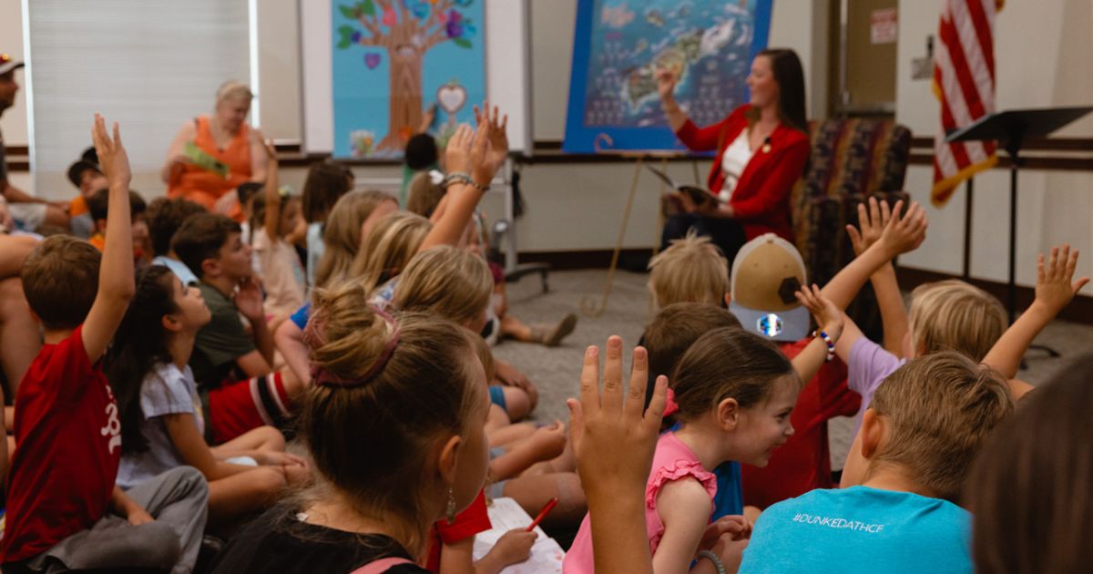 This image was taken at a 'See You at the Library' event in August of 2023. The image shows a group of children sitting in front of a woman reading them a book during story hour at the library.