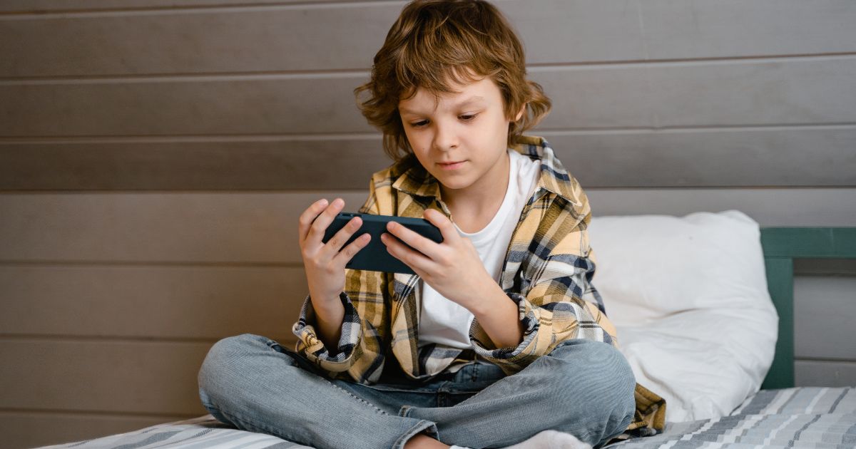 This image shows a young boy sitting on the bed while using his mobile phone.