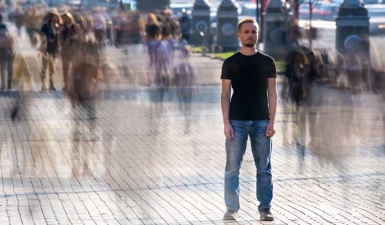 This image shows a young man standing still while many blurred people pass by him on the sidewalk.