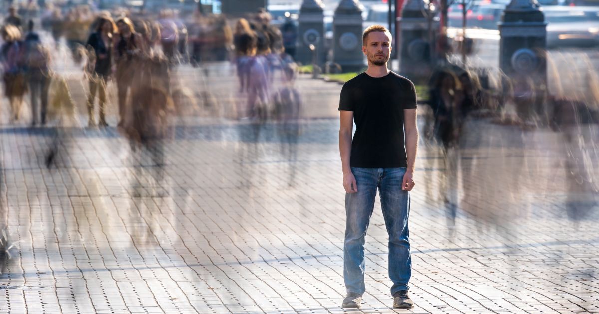 This image shows a young man standing still while many blurred people pass by him on the sidewalk.