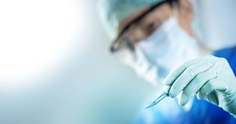 A surgeon's hand holds a scalpel with his face blurred in the background.