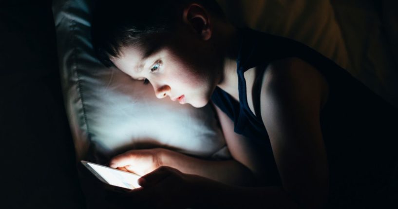 A young boy looks at a tablet or smartphone at night in bed.