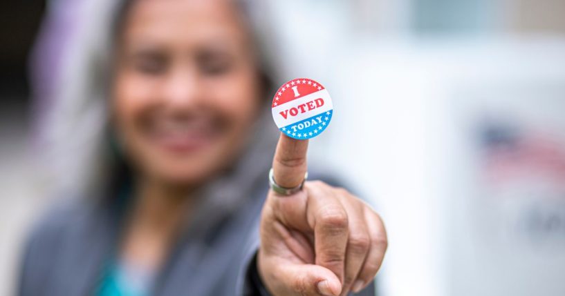 This image shows a close-up of an "I voted sticker" that a senior woman is holding up. Her face is blurred.