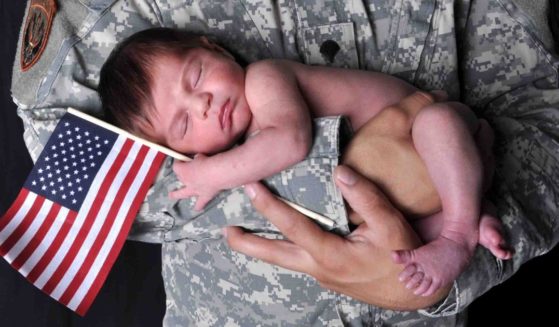 This image shows an American soldier holding a newborn baby who is holding onto an American flag while sleeping.