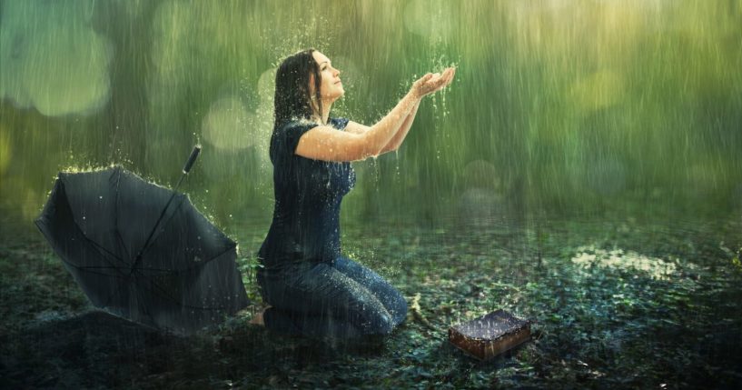 This image shows a woman kneeling in the rain with an umbrella behind her and a book in front of her on the ground.