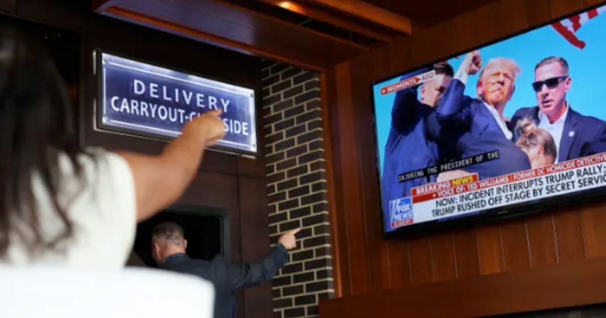 This image shows a young woman pointing at a television in a restaurant that is showing former President Trump shortly after being shot at his Pennsylvania rally on July 14.