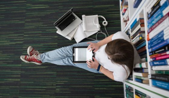 This image shows a female student sitting on the floor in the library and leaning against a bookshelf. She is using a tablet and appears to be studying.