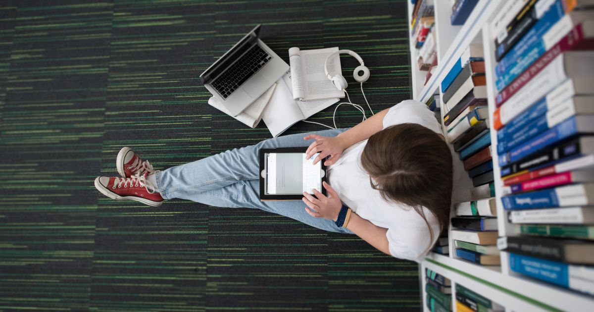 This image shows a female student sitting on the floor in the library and leaning against a bookshelf. She is using a tablet and appears to be studying.