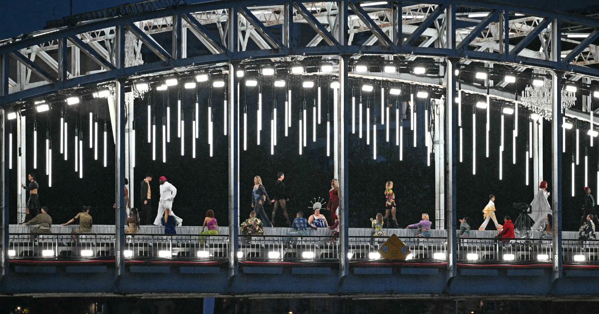 Models present creations while walking a catwalk erected along the Passerelle Debilly bridge as the boat carrying the delegations of Romania and Rwanda sails underneath along the Seine river during the opening ceremony of the Paris 2024 Olympic Games in Paris on July 26, 2024.