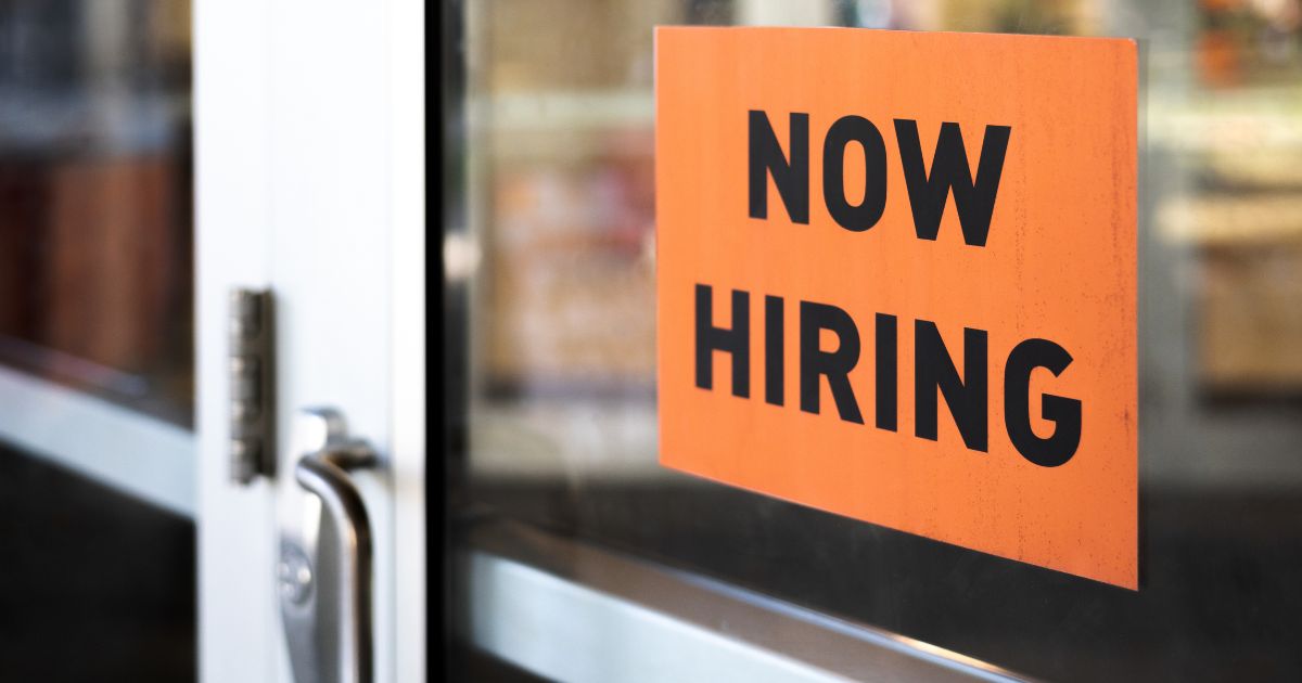 This stock image shows a “Now Hiring” sign hanging on the door of a business.