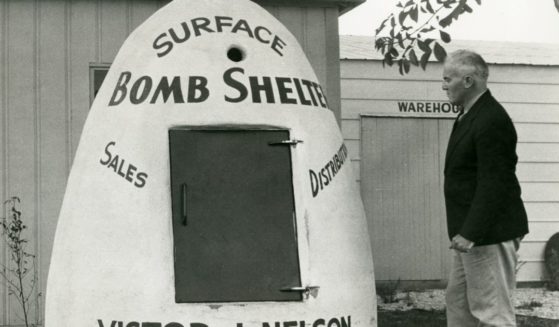 A black-and-white photo shows a man standing next to a surface bomb shelter that is advertising Victor J. Nelson's sales and distribution business in 1951.