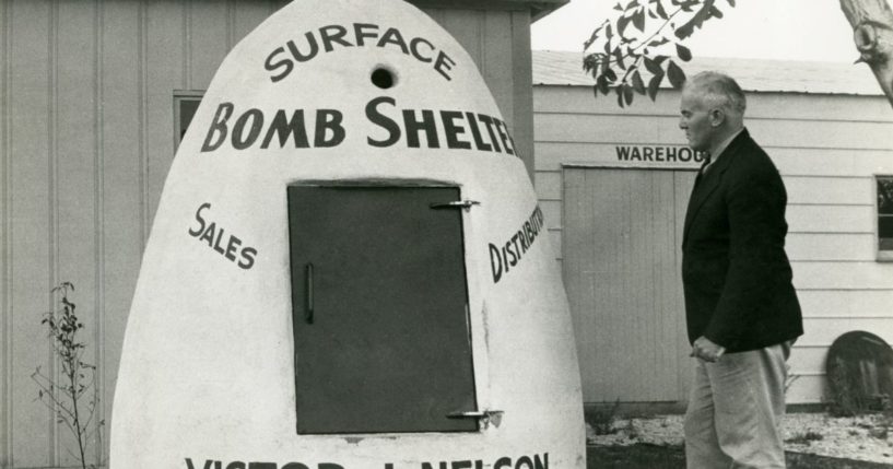 A black-and-white photo shows a man standing next to a surface bomb shelter that is advertising Victor J. Nelson's sales and distribution business in 1951.