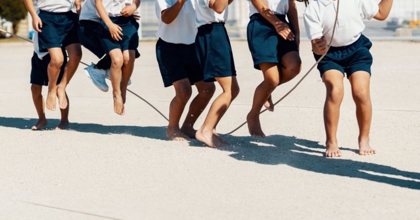 A class of young children practice jump roping, barefoot, with their teacher.