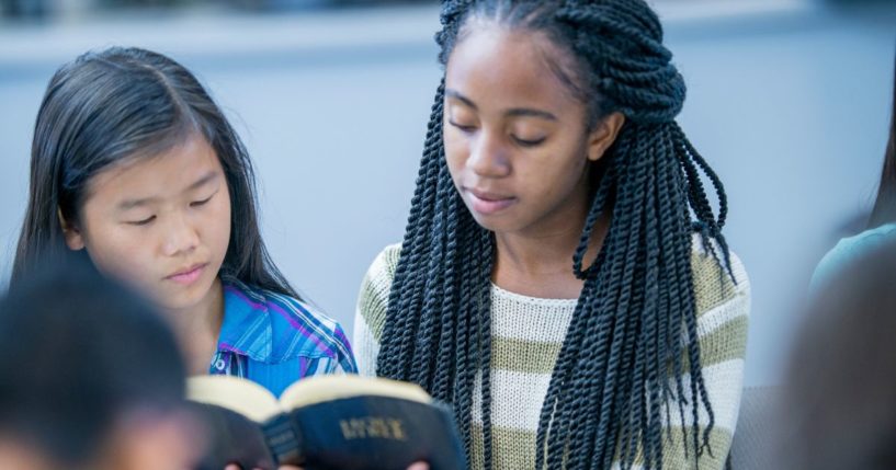 A high school age girl is holding the Bible and reading it with her fellow classmates during a Bible study.